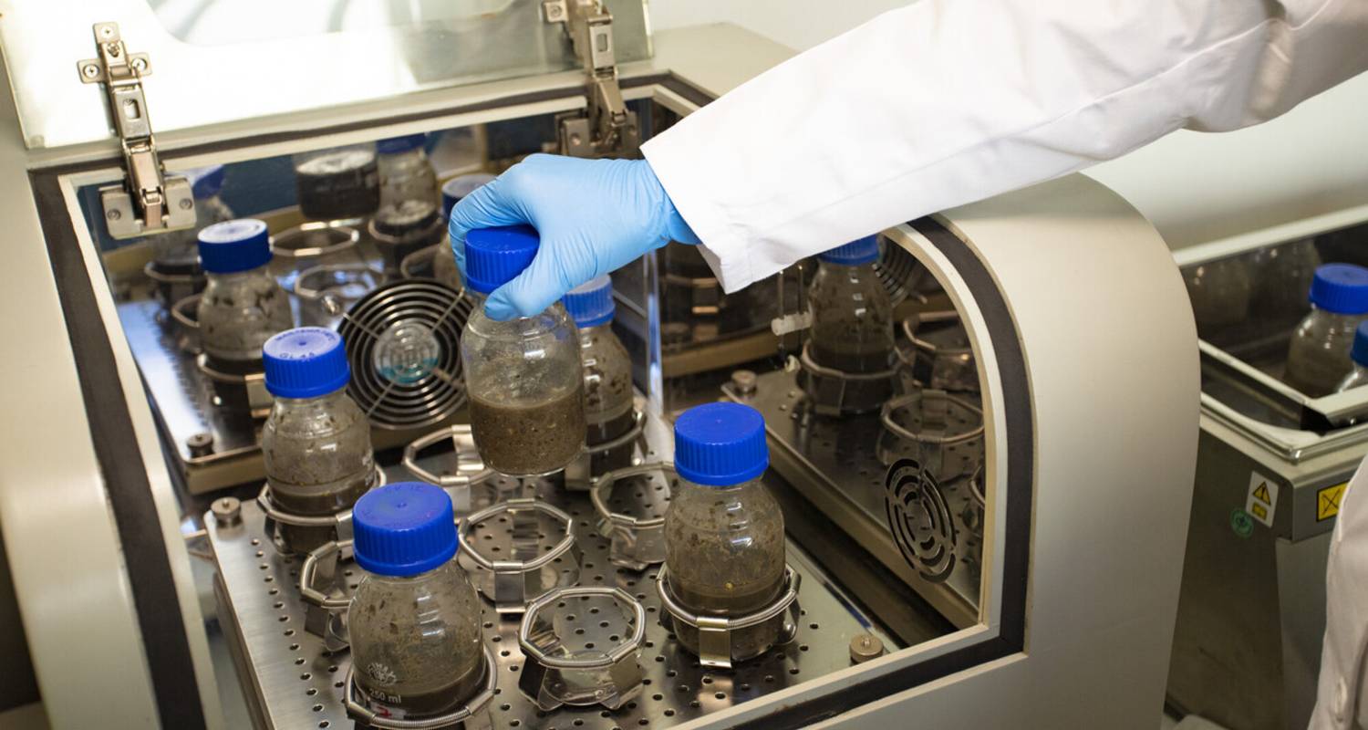 Mano con guante colocando una botella con una muestra en una máquina del laboratorio.